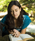 Young woman reading her Bible by a stream in summer.