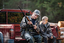 Father and son sitting in a pickup truck after hunting in forest with the dad showing the boy the mechanism of a shotgun rifle.